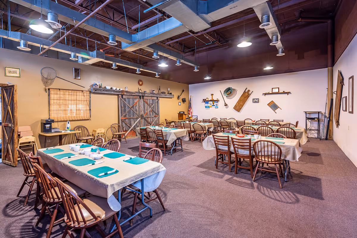 A spacious dining room with multiple tables covered in beige tablecloths and green placemats, surrounded by wooden chairs. The room has a rustic decor with fishing nets, wooden shelves, and various vintage items on the walls. The ceiling features exposed beams and industrial lighting.