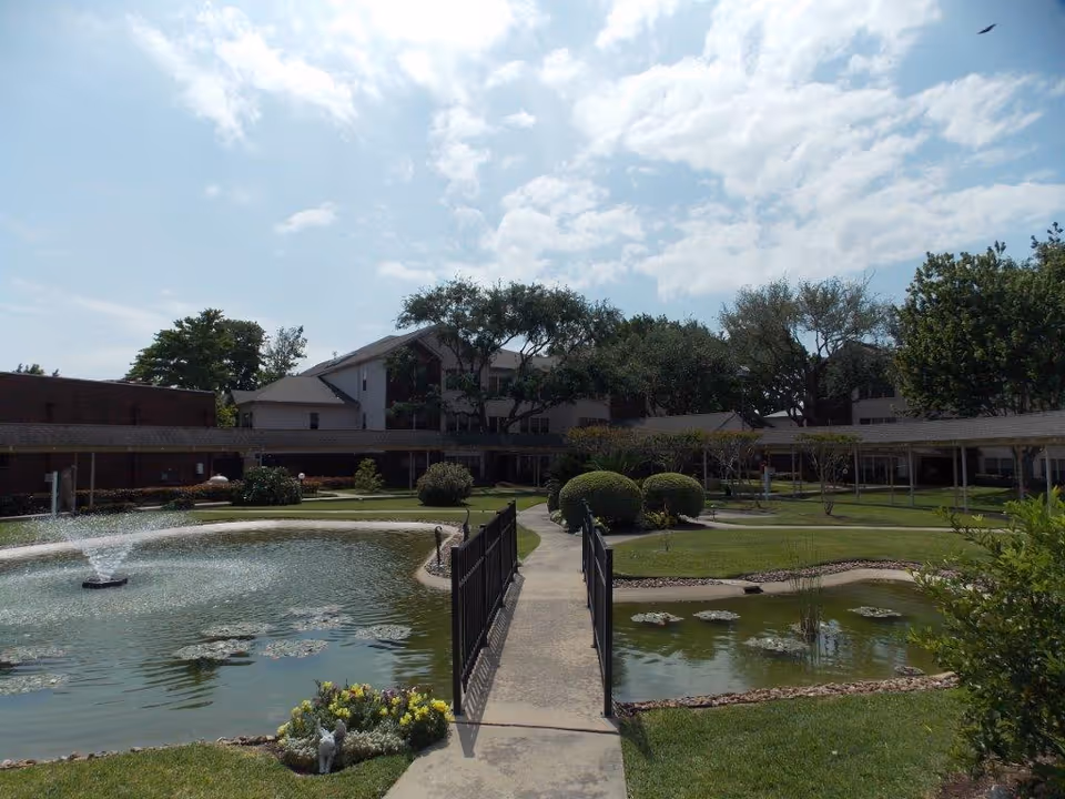 Outdoor view of Lone Star Living facility showing a landscaped garden with a pond featuring a water fountain, a concrete walkway with a small bridge over the pond, trimmed bushes, trees, and a multi-story building in the background under a partly cloudy sky.