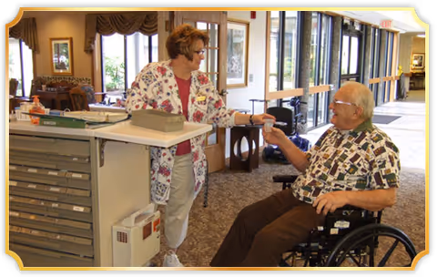 A healthcare worker in a floral scrub top hands a small cup to an elderly man sitting in a wheelchair inside a well-lit facility hallway with large windows.