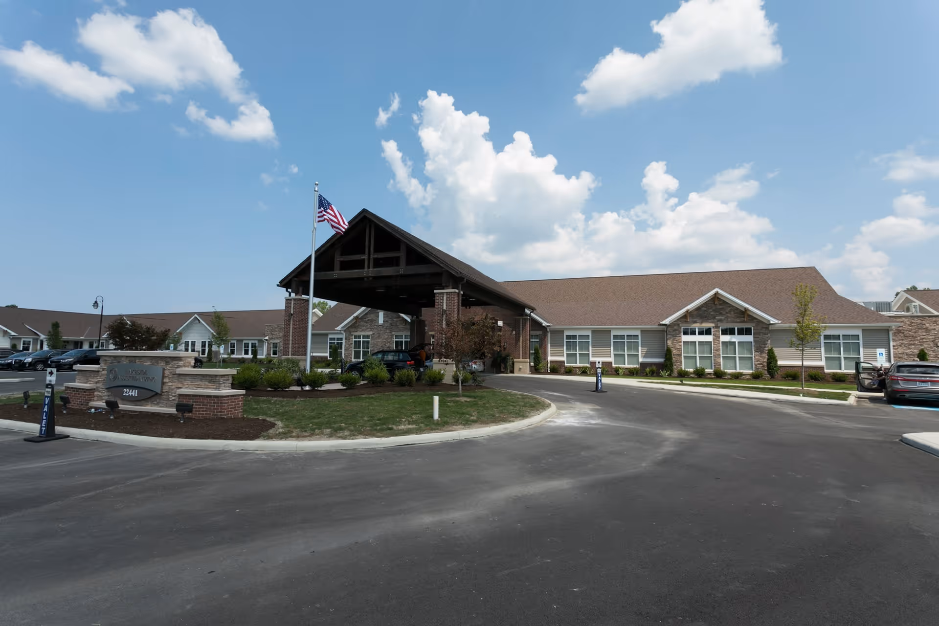 Exterior view of a single-story senior living facility building with a covered entrance, an American flag on a flagpole, landscaped greenery, and a parking area under a partly cloudy sky.
