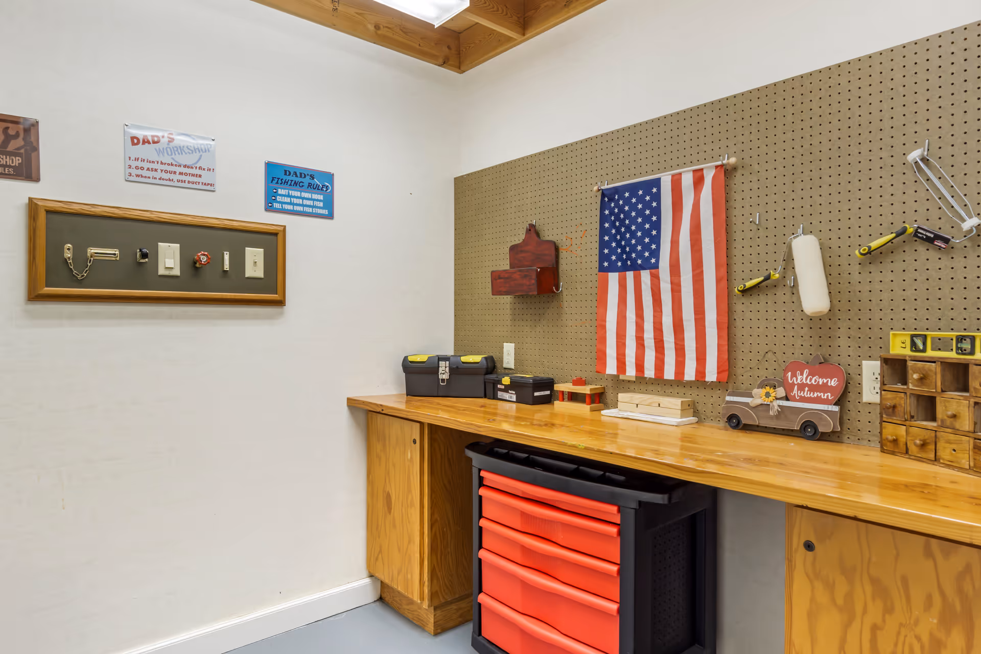 A workshop area with a wooden workbench, pegboard wall holding tools and an American flag. There are small decorative signs on the wall, including one that says 'Welcome Autumn' and another with 'Dad's Fishing Rules'. A red tool chest with multiple drawers is under the workbench.