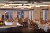 A spacious dining room with multiple wooden tables and chairs arranged neatly. The tables are covered with white tablecloths and set with glassware and napkins. Large windows allow natural light to fill the room, and ceiling lights provide additional illumination. The room has a warm and inviting atmosphere.