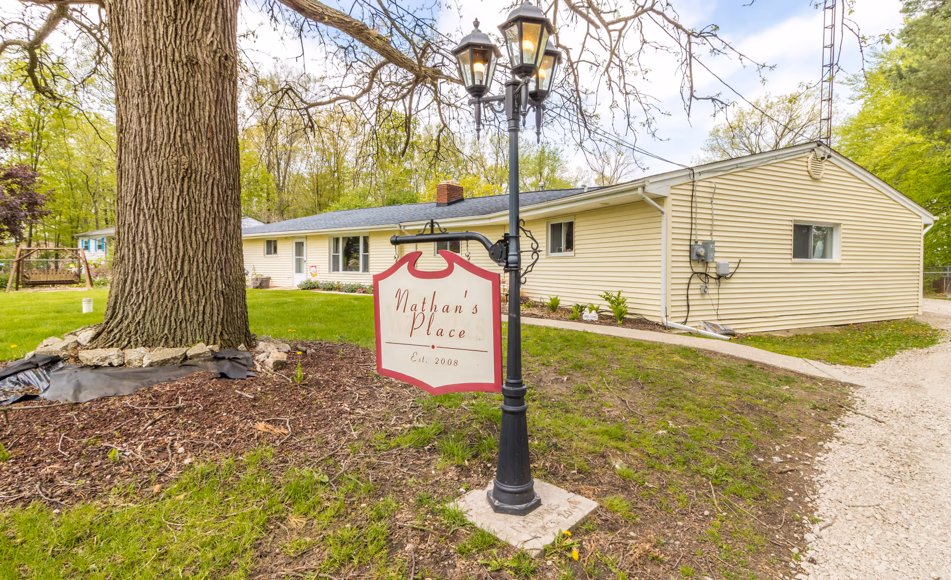Front exterior of a single-story beige building with a lawn, large tree, and a lamp-post sign reading "Nathan's Place".