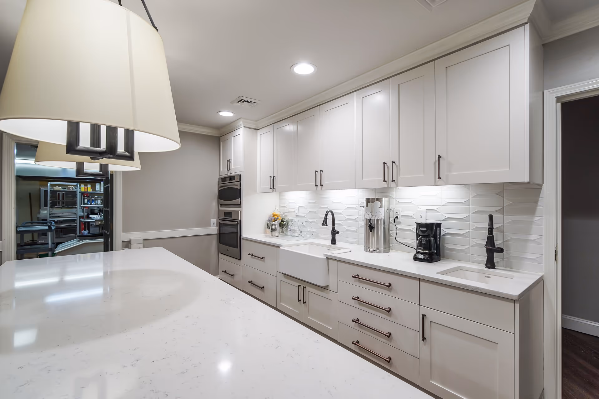 Modern kitchen area with white cabinets, two black faucets, a coffee maker, and a large white countertop island. The backsplash features a textured white tile design, and there are built-in ovens on the left side. Pendant lights hang above the island, and a doorway leads to a storage or pantry area.