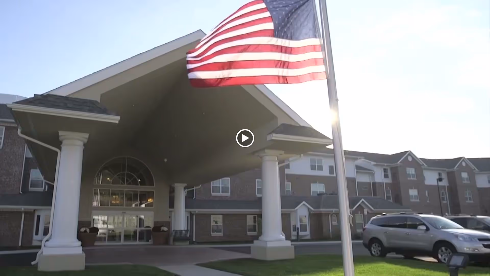 Front exterior view of Virginia Greens Retirement Community building with a covered entrance supported by white columns, an American flag on a flagpole in the foreground, and parked cars to the right.