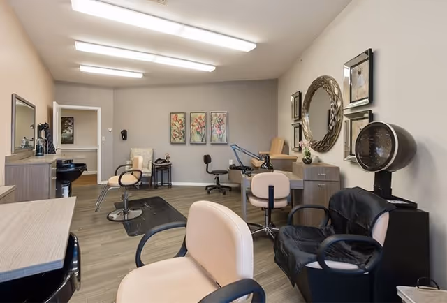 Interior view of a hair salon area with multiple styling chairs, a hair dryer chair, mirrors, and framed floral artwork on the walls. The room has wood flooring and neutral-colored walls with overhead fluorescent lighting.