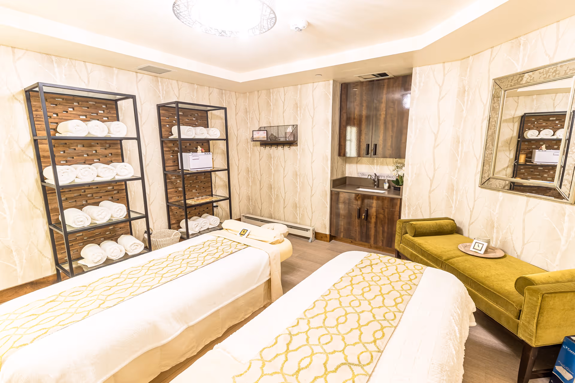 A bright and cozy spa room with two massage tables covered in white linens and yellow patterned runners. The room features two black metal shelves stocked with rolled white towels, a small wooden cabinet with a sink, and a green upholstered bench with a small tray and framed picture on it. The walls have a light beige wallpaper with a subtle tree branch pattern, and a large decorative mirror hangs on one wall.