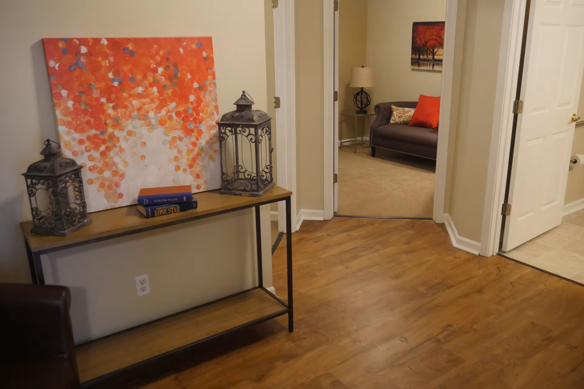 Interior view of a hallway with wooden flooring leading to a carpeted room with a dark brown sofa adorned with red and patterned pillows. A side table with a lamp and a painting of red trees is visible in the room. In the hallway, there is a console table with two decorative lanterns, three books, and a large abstract painting with red, orange, and white colors leaning against the wall.