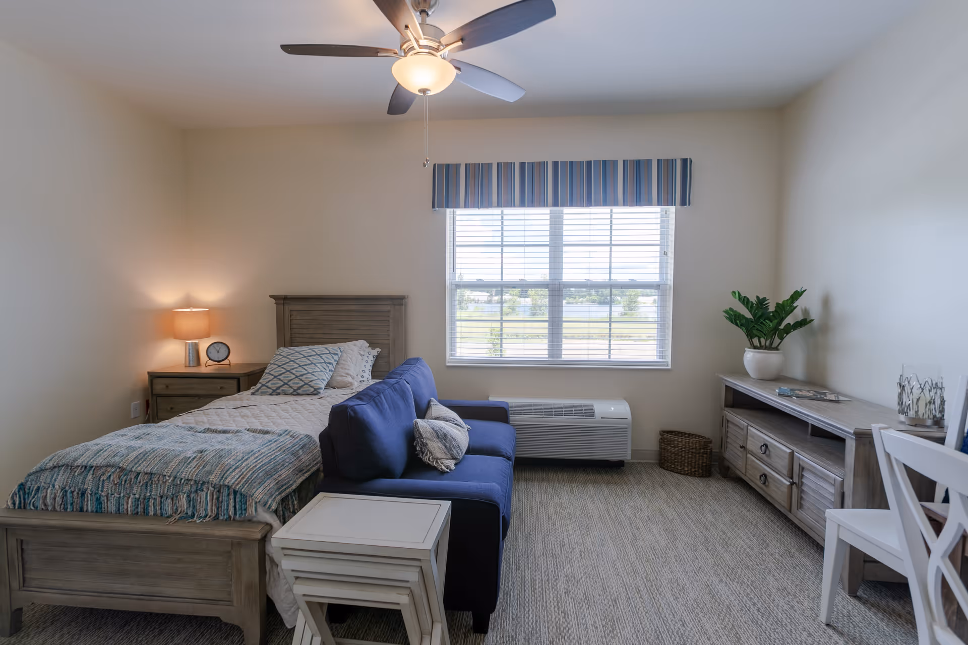 A cozy senior living bedroom featuring a single bed with a wooden headboard and a blue patterned blanket. Next to the bed is a wooden nightstand with a lamp and a clock. In front of the bed is a small blue sofa with a cushion. The room has a large window with blinds and a striped valance, letting in natural light. There is a wooden dresser with a potted plant and decorative items, and a white chair is partially visible on the right side. The ceiling has a fan with a light fixture.