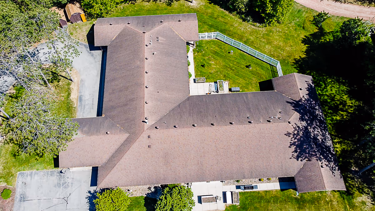 Aerial view of a single-story building with a brown roof surrounded by green grass and trees. The building has a fenced grassy area on one side and paved walkways around it.