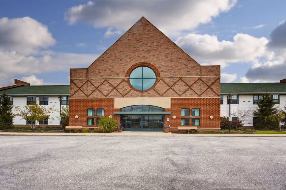 Front exterior view of Stratford Commons Assisted Living facility featuring a large brick building with a triangular roof section, a circular window above the main entrance, and two benches on either side of the entrance. The sky is partly cloudy.