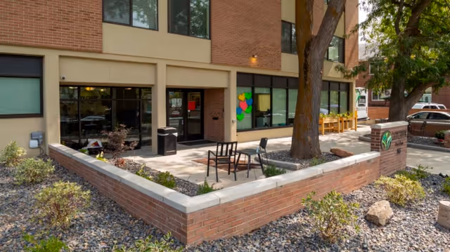 Front exterior of a brick senior living facility showing the entrance, a small patio with chairs, and landscaping.