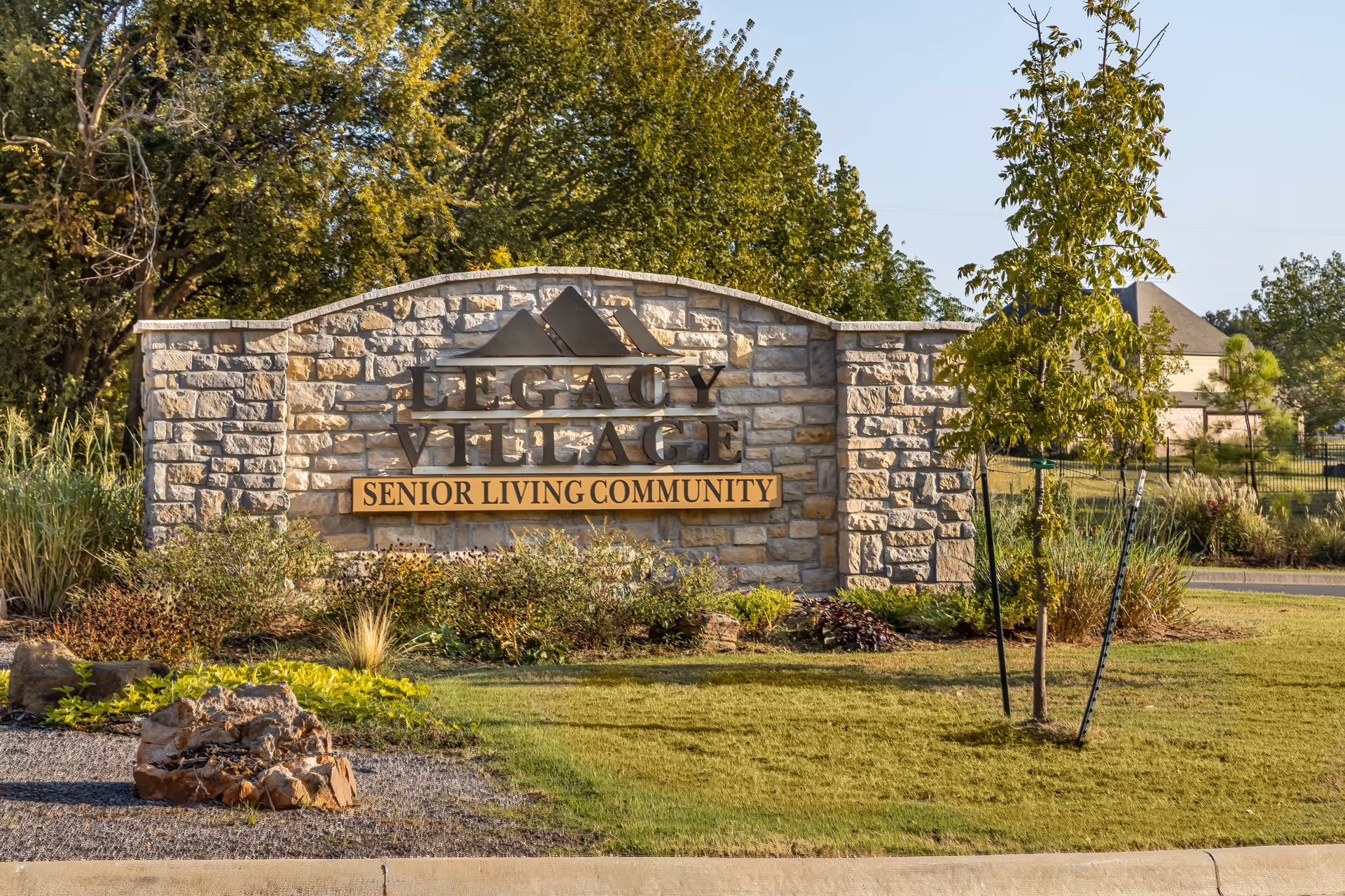 Stone entrance sign for Legacy Village Senior Living Community surrounded by landscaped greenery and trees under a clear sky.
