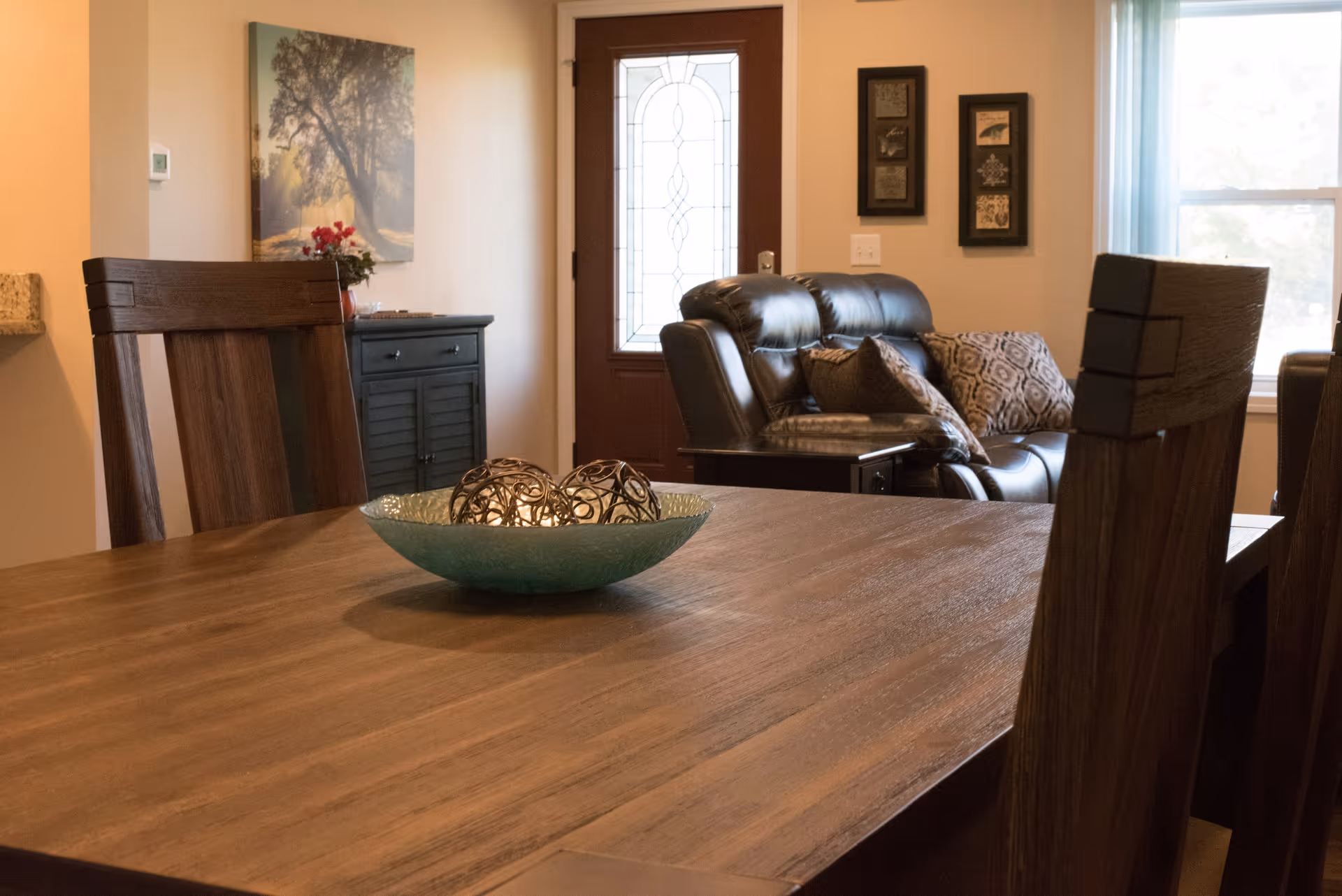 Interior view of a living and dining area with a wooden dining table and chairs in the foreground, a decorative bowl with ornamental balls on the table, a leather sofa with patterned cushions near a window, a wooden cabinet with a flower vase, and a door with a decorative glass panel.