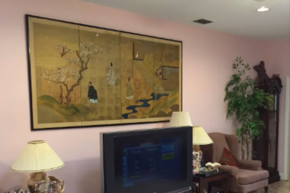 A cozy living room with pink walls featuring a large framed artwork depicting traditional Asian figures and scenery. The room contains a television, two table lamps with cream-colored shades, a comfortable armchair with a decorative pillow, a tall green plant, and a tall wooden grandfather clock in the corner.