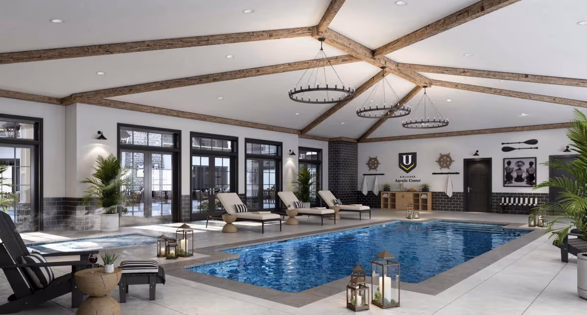 Indoor swimming pool area with lounge chairs, potted plants, and decorative lanterns. The ceiling has exposed wooden beams and circular chandeliers. Large windows and doors line the walls, allowing natural light to enter. Nautical-themed decorations and a sign reading 'Wagner Aquatic Center' are visible on the far wall.