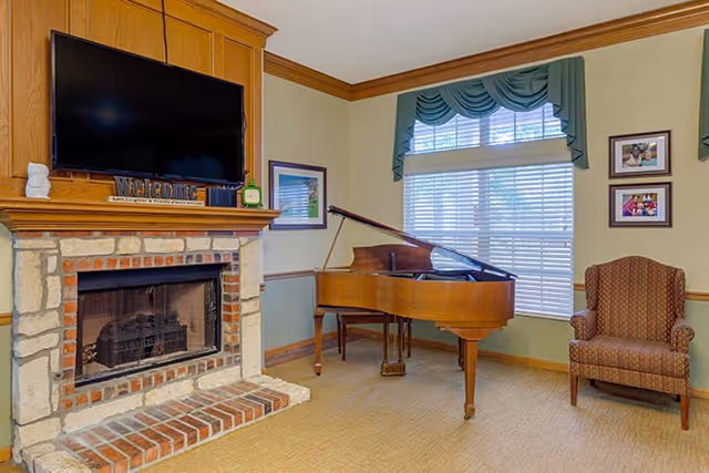 A cozy living room area featuring a stone and brick fireplace with a mounted flat-screen TV above it. On the mantel, there is a decorative 'welcome' sign and a small clock. To the right of the fireplace, there is a wooden grand piano and a patterned upholstered armchair. The room has a large window with white blinds and green valances, and framed pictures hang on the wall.