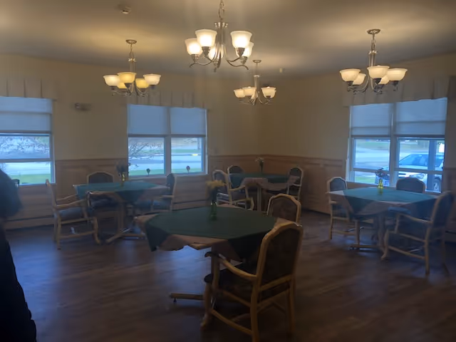 A dining room with several square tables covered with green tablecloths, each surrounded by wooden chairs with cushioned seats and backs. Small flower vases are placed on each table. The room has large windows with white blinds, wooden paneling on the lower half of the walls, and multiple ceiling light fixtures with frosted glass shades.