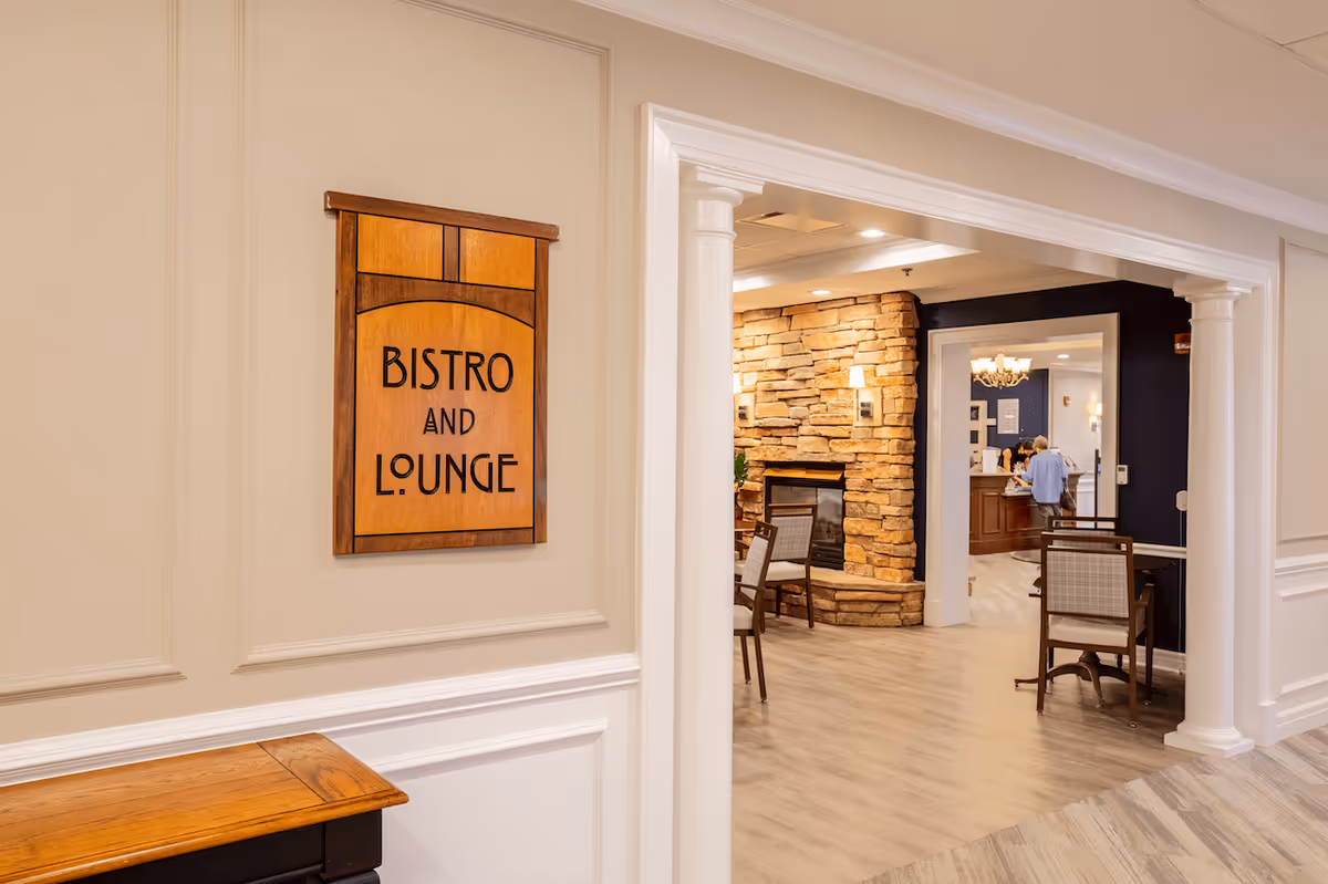 Interior view of a senior living facility showing an entrance to a bistro and lounge area. The entrance is framed by white columns and molding, with a wooden sign on the wall reading 'Bistro and Lounge'. Inside, there are chairs and tables near a stone fireplace, and a reception desk with a person standing behind it.