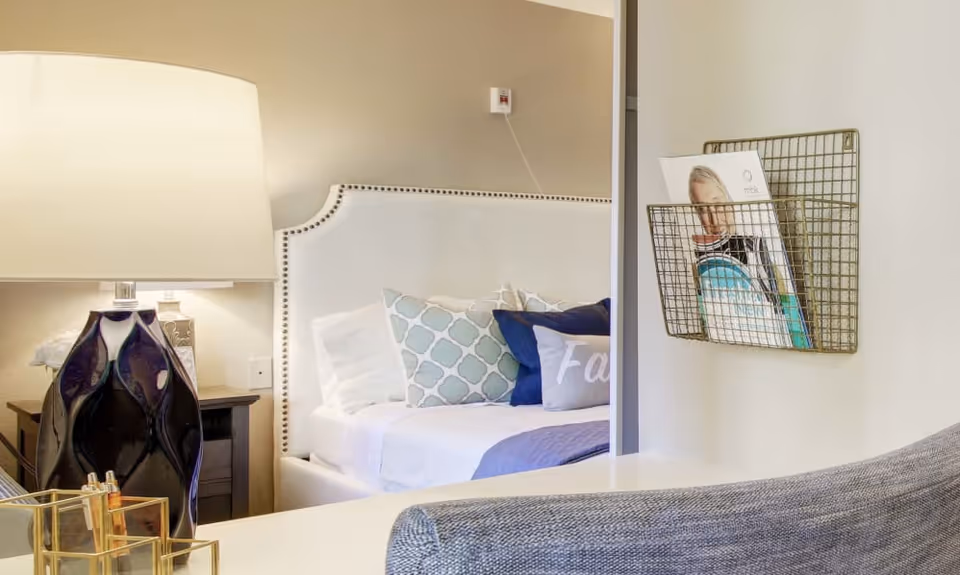 A cozy bedroom corner with a white upholstered headboard adorned with nailhead trim, several decorative pillows in blue and white patterns, a bedside table with a dark blue ceramic lamp, and a wire wall-mounted magazine holder containing brochures. A gray upholstered chair is partially visible in the foreground.
