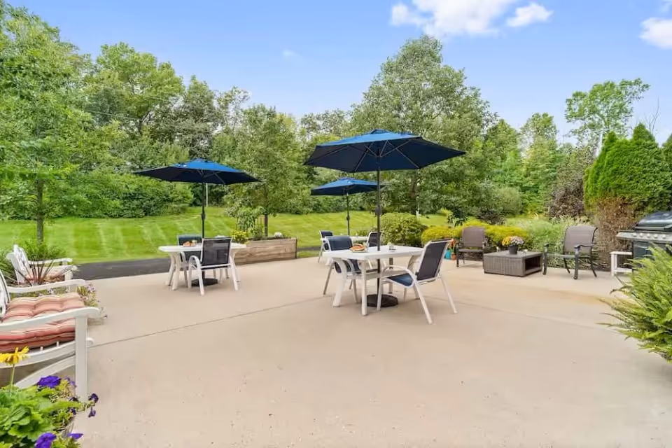 Outdoor patio area with several tables and chairs, each table shaded by a blue umbrella. The patio is surrounded by green trees and bushes under a clear blue sky.