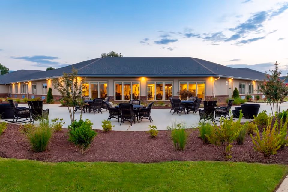 Outdoor patio area of Whitetail Springs Memory Care Community with multiple black wicker chairs and tables arranged on a concrete surface, surrounded by landscaped bushes and plants, with the building in the background illuminated by exterior lights during dusk.