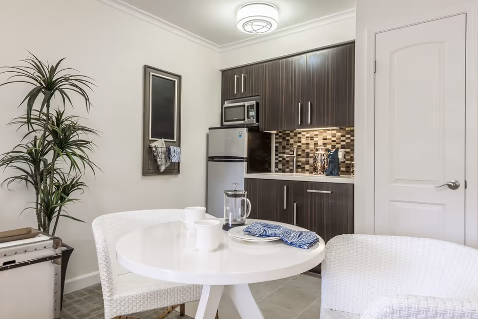Small kitchenette with dark wood cabinets, a compact refrigerator and a round white dining table with wicker chairs.