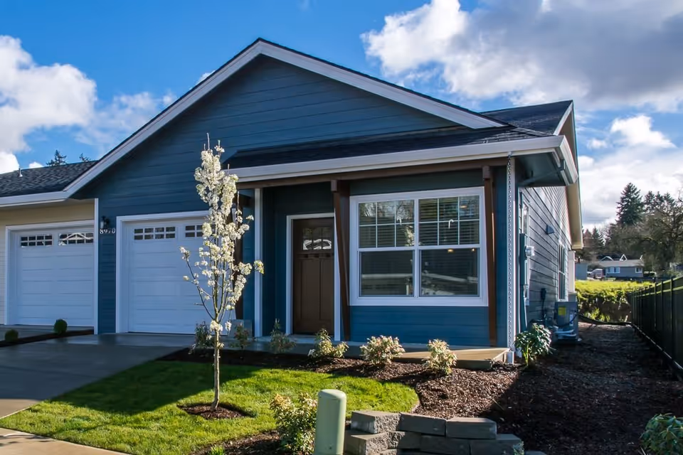 Exterior view of a single-story blue house with white trim, a front porch, a garage with two white doors, a small tree with white blossoms, and a well-maintained lawn under a partly cloudy sky.