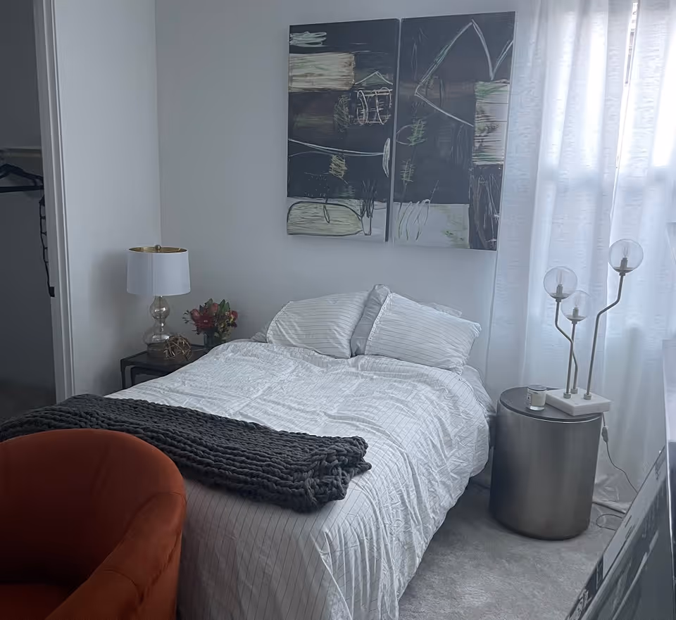 A cozy bedroom with a neatly made bed featuring white striped bedding and two pillows. Above the bed, there are two abstract paintings. To the left of the bed is a small nightstand with a clear glass lamp and a small bouquet of flowers. To the right, there is a round metallic side table with a modern lamp with three glass globes and a small candle. A burnt orange armchair is partially visible in the foreground. The room has light-colored walls and a window with sheer white curtains allowing natural light to enter.