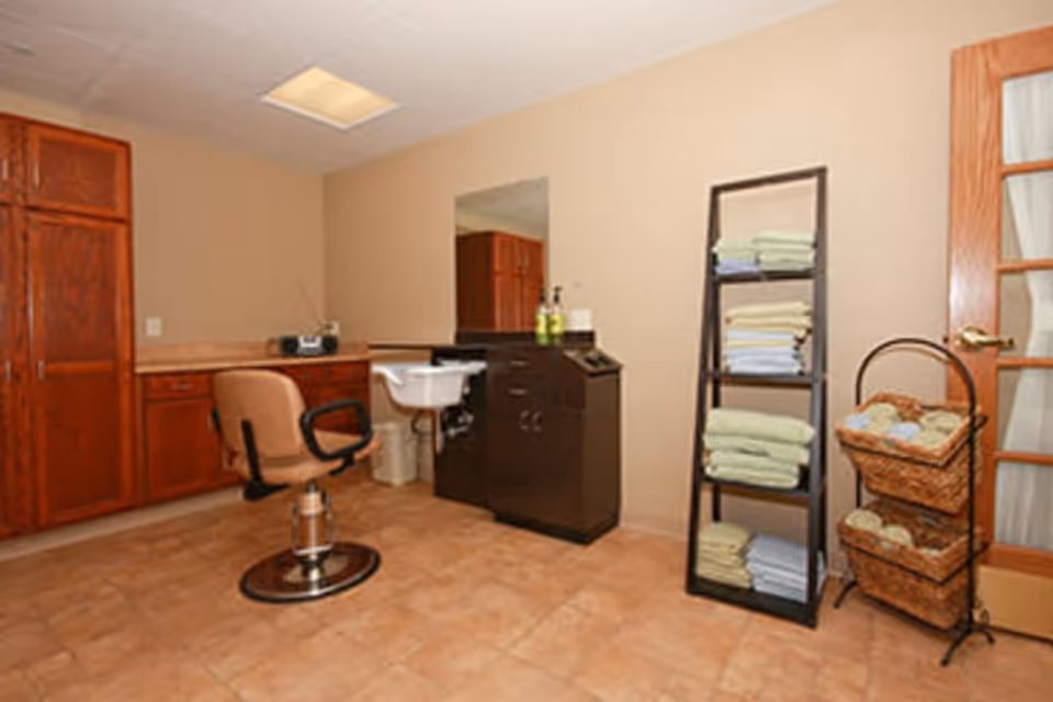 Interior of a senior living facility room with a salon chair in front of a small sink and mirror. There are wooden cabinets along one wall, a shelving unit with neatly folded towels, and a basket stand with additional towels. The room has beige walls and tiled flooring.