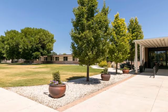 Outdoor view of Parke View Rehabilitation & Care Center showing a landscaped area with green grass, several trees, large potted plants with flowers, and a paved walkway leading to the building entrance with a covered porch.