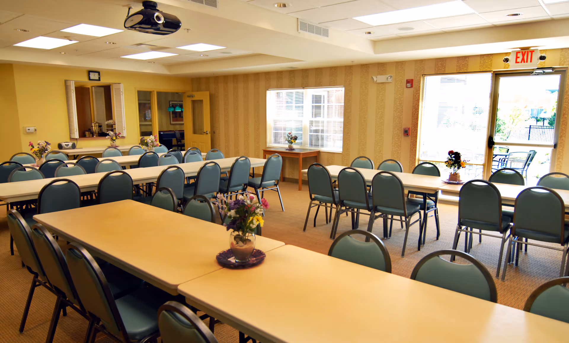 A well-lit dining room with multiple long tables and blue cushioned chairs arranged around them. Each table has a small vase with flowers. There is a window letting in natural light and a door with an exit sign above it leading outside to a patio area with outdoor seating. The walls have a patterned wallpaper and the ceiling has recessed lighting and a mounted projector.