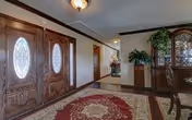 Interior view of a hallway in an assisted living facility featuring double wooden doors with oval glass panels, a patterned area rug on the floor, and a wooden cabinet with glass doors and plants on top along the wall.