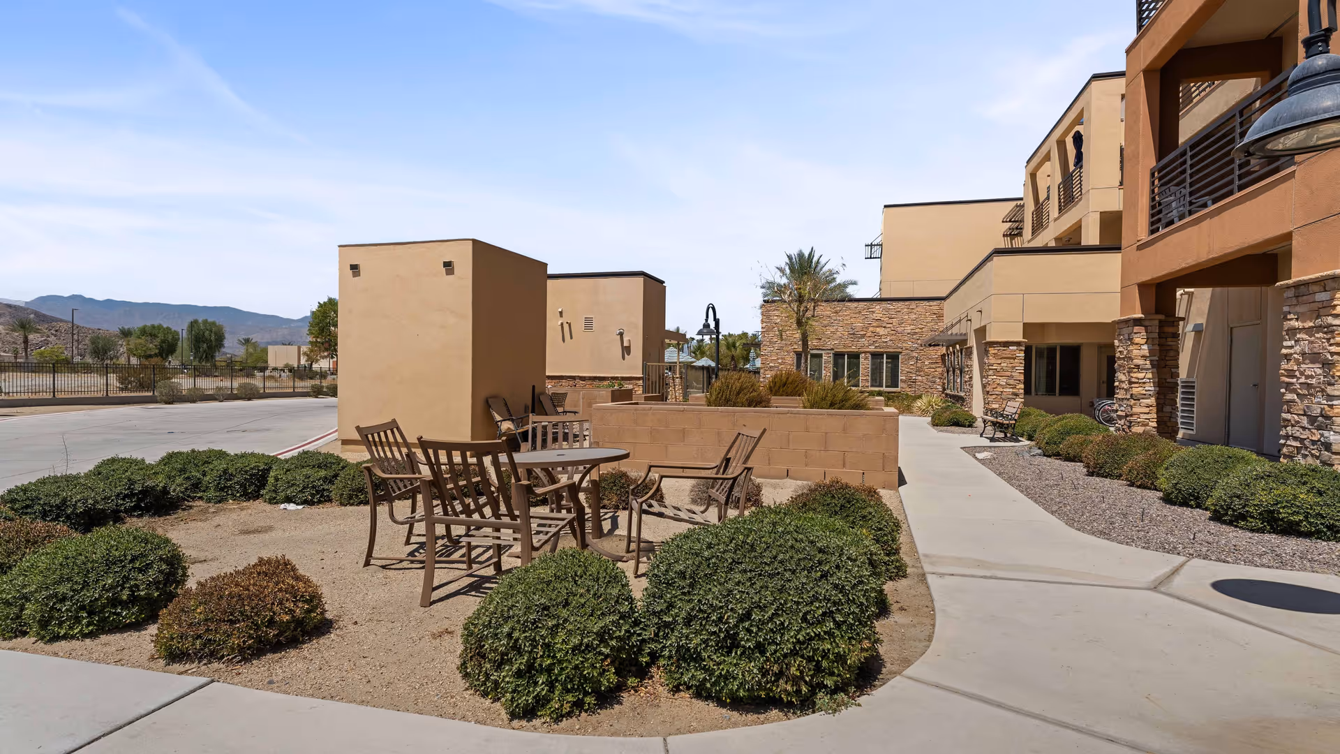 Outdoor patio area at The Palms At La Quinta Gracious Retirement Living featuring a round table with four chairs surrounded by small bushes and desert landscaping. The building exterior has stone and stucco finishes with balconies and windows. Mountains and clear sky are visible in the background.
