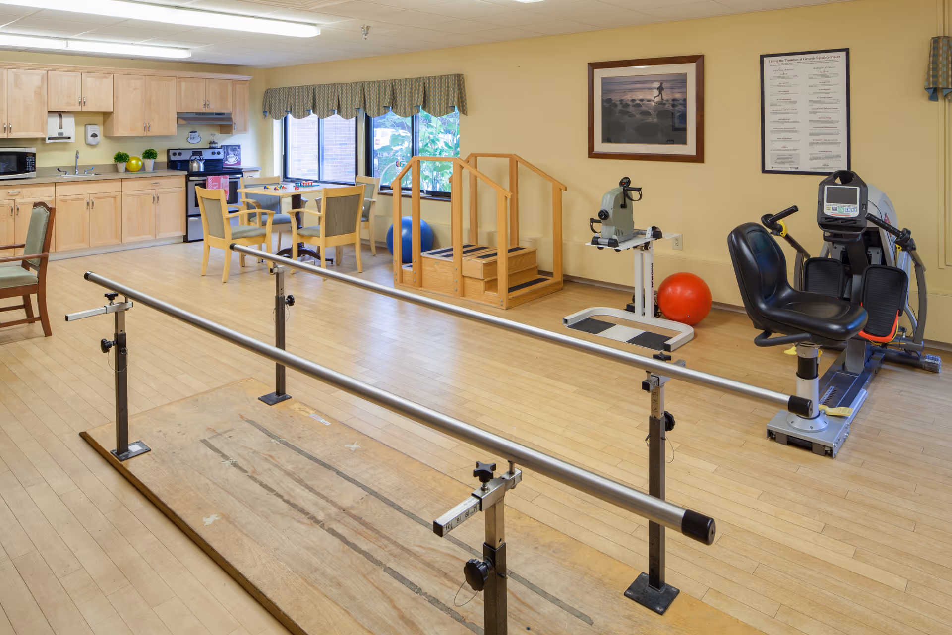 Rehabilitation therapy room featuring parallel walking bars, exercise equipment, a table with chairs, and a kitchenette.