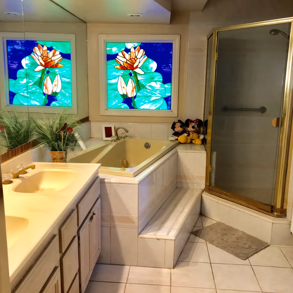 A bathroom with a beige bathtub surrounded by tiled steps, a glass-enclosed shower with a gold frame, a double sink vanity with a large mirror, a potted plant on the counter, and a stained glass window featuring a floral design. Two plush Mickey and Minnie Mouse toys are placed on the ledge next to the bathtub.