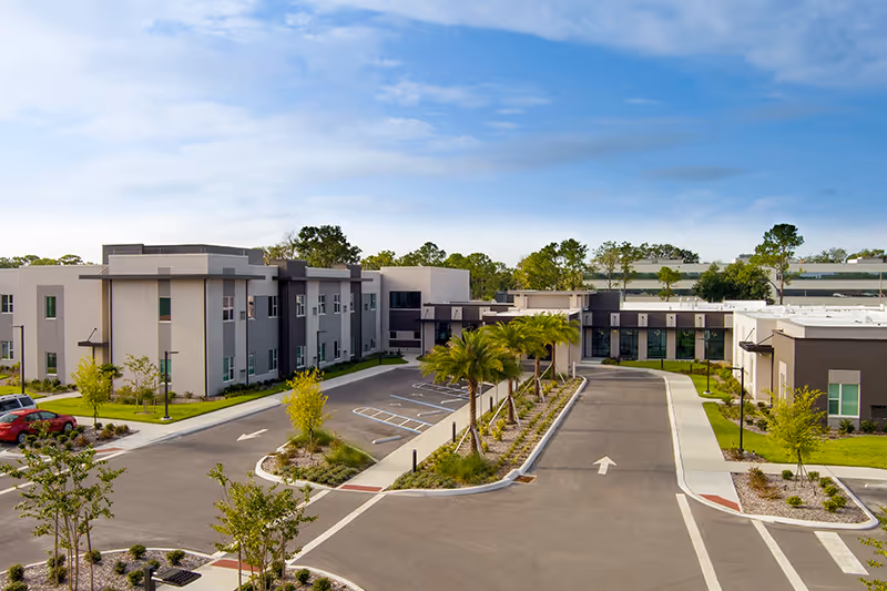 Exterior view of the Village on the Green showing the entrance driveway, landscaped median with palm trees, and modern low-rise buildings under a blue sky.