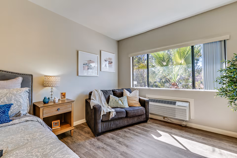 A bright senior living bedroom with a bed on the left side featuring patterned bedding and pillows. Next to the bed is a wooden nightstand with a lamp, small decorative items, and framed photos. A dark upholstered loveseat with several throw pillows and a blanket is positioned near a large window with vertical blinds, allowing natural light to fill the room. A green plant is partially visible on the right side, and the floor is covered with wood-style flooring.