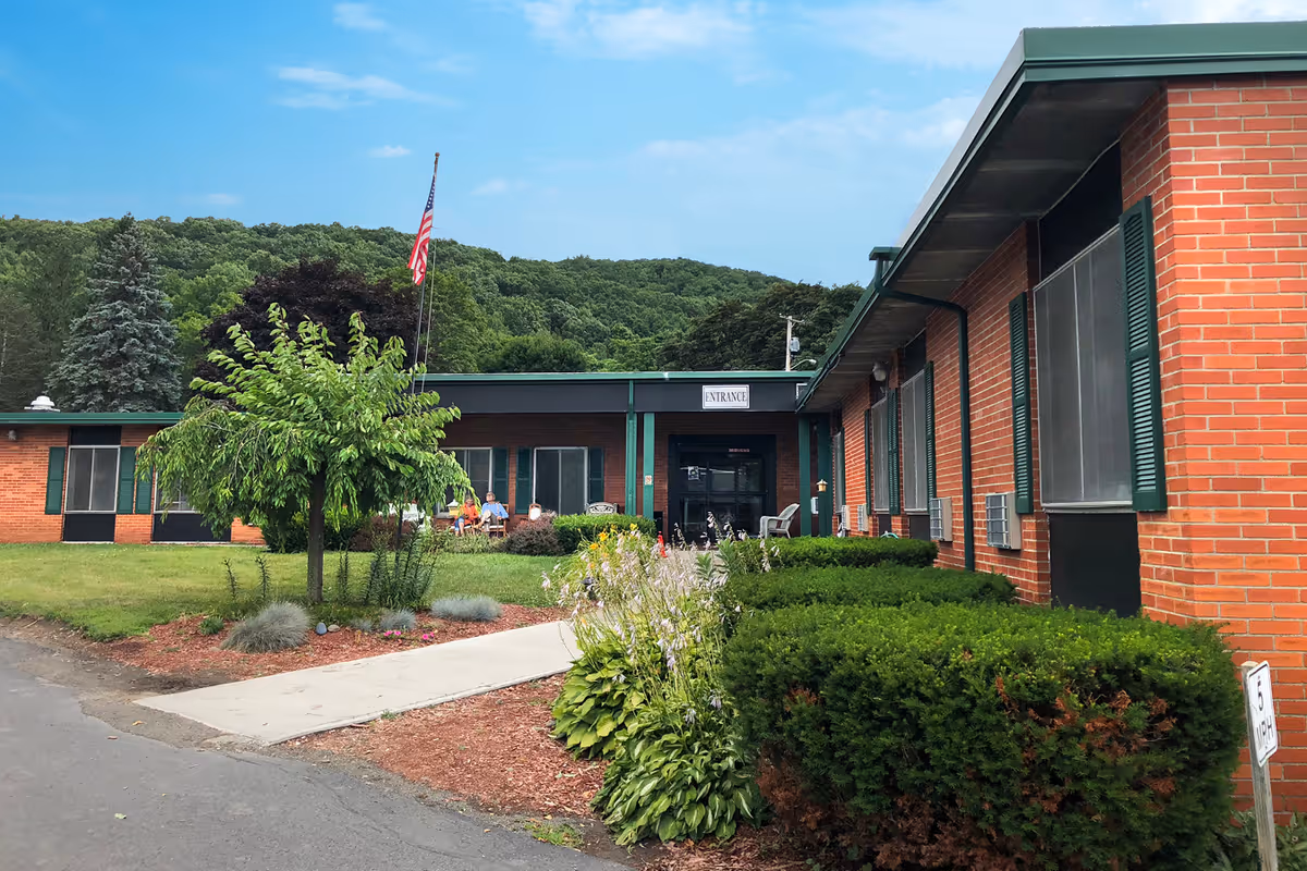 Exterior view of Chestnut Park Rehabilitation and Nursing Center showing a single-story brick building with green shutters and a green roof. There is a sidewalk leading to the entrance, an American flag on a flagpole, some bushes and small trees, and a few people sitting outside near the entrance. The background features a forested hillside under a blue sky with some clouds.