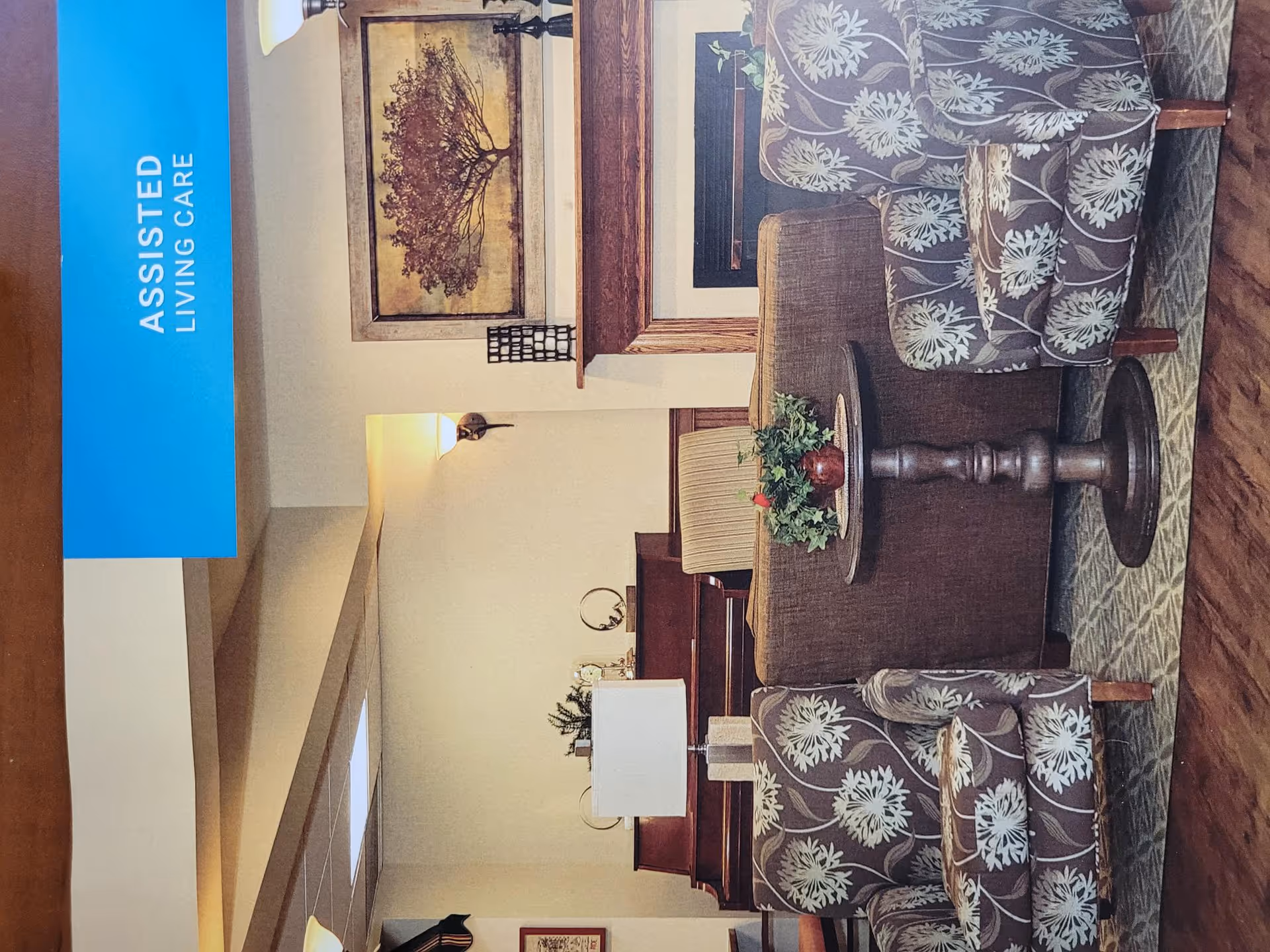A cozy assisted living care lounge area with two floral patterned armchairs, a brown sofa, a round wooden table with a plant centerpiece, a fireplace with a framed tree artwork above it, and a piano in the background.