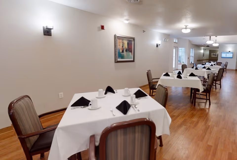 A dining room with several tables covered in white tablecloths, each set with black folded napkins, white cups, saucers, and silverware. The room has wooden flooring, beige walls with modern wall sconces, and framed artwork. Chairs with wooden frames and striped upholstery surround the tables. The room is well-lit with ceiling lights and has windows and doors along one side.