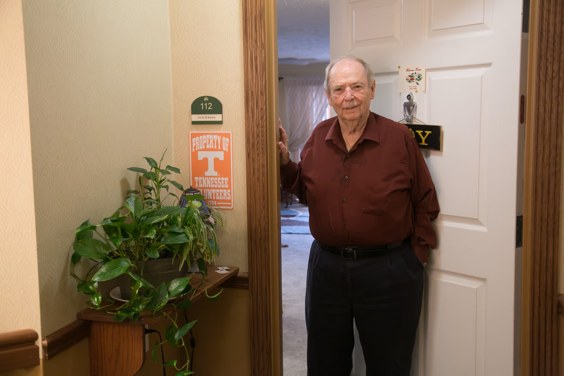 An elderly man wearing a maroon shirt and dark pants stands in the doorway of a room numbered 112 at Sherrill Hills Retirement Resort. There is a green plant on a wooden shelf next to the door, and a sign on the wall reads 'Property of Tennessee Volunteers'.