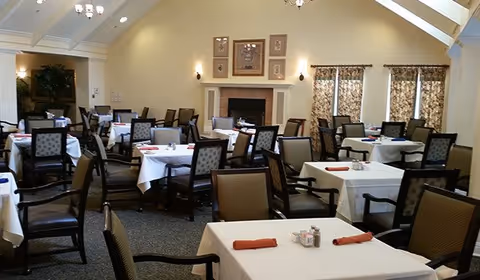 Dining room with multiple tables covered with white tablecloths, each set with folded orange napkins and condiments. The room has high ceilings with exposed beams, a fireplace with framed artwork above it, patterned curtains on the windows, and several upholstered chairs around the tables.