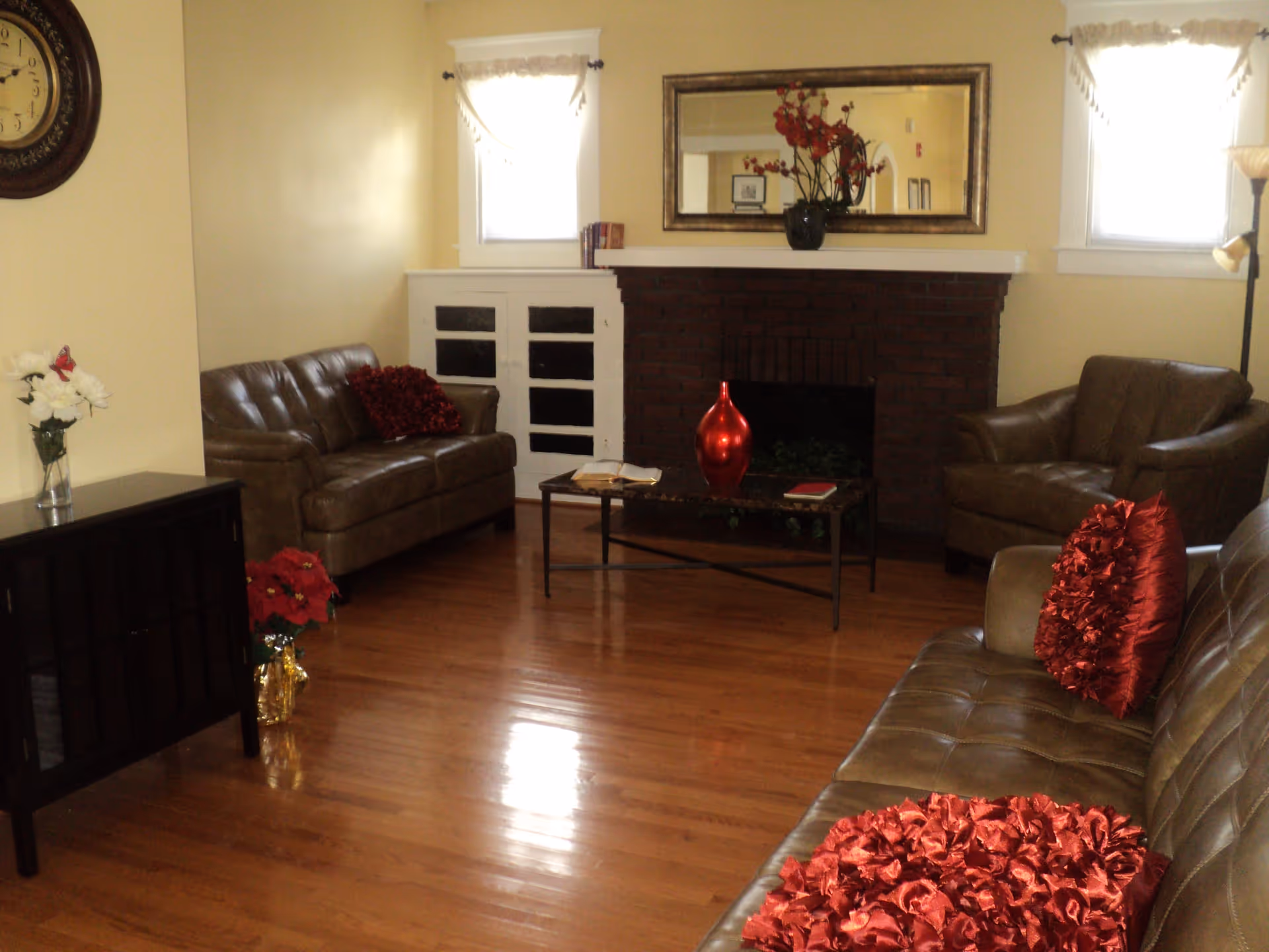 A cozy living room with three brown leather sofas adorned with red decorative pillows. The room features a wooden floor, a brick fireplace with a large mirror above it, and a coffee table with a red vase and books. There are two small windows with white curtains on either side of the fireplace, a wall clock, a floor lamp, and a small cabinet with flowers on top.