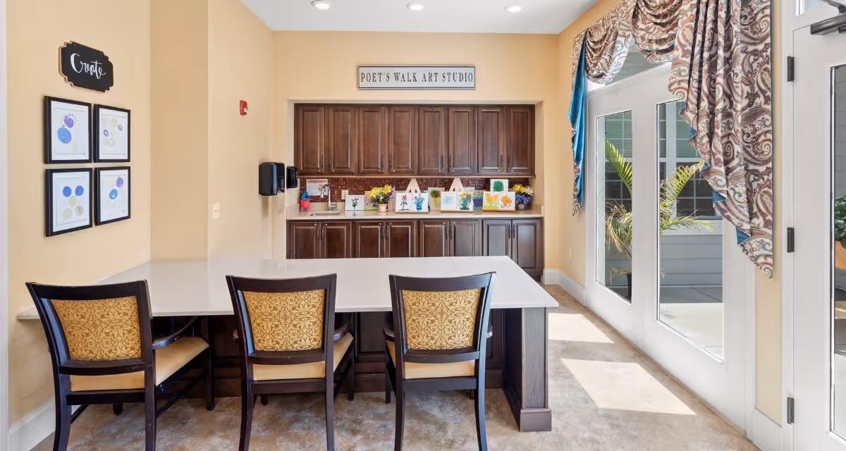 Bright art studio interior in a memory-care community with a large island and three chairs facing wooden cabinets and glass doors.
