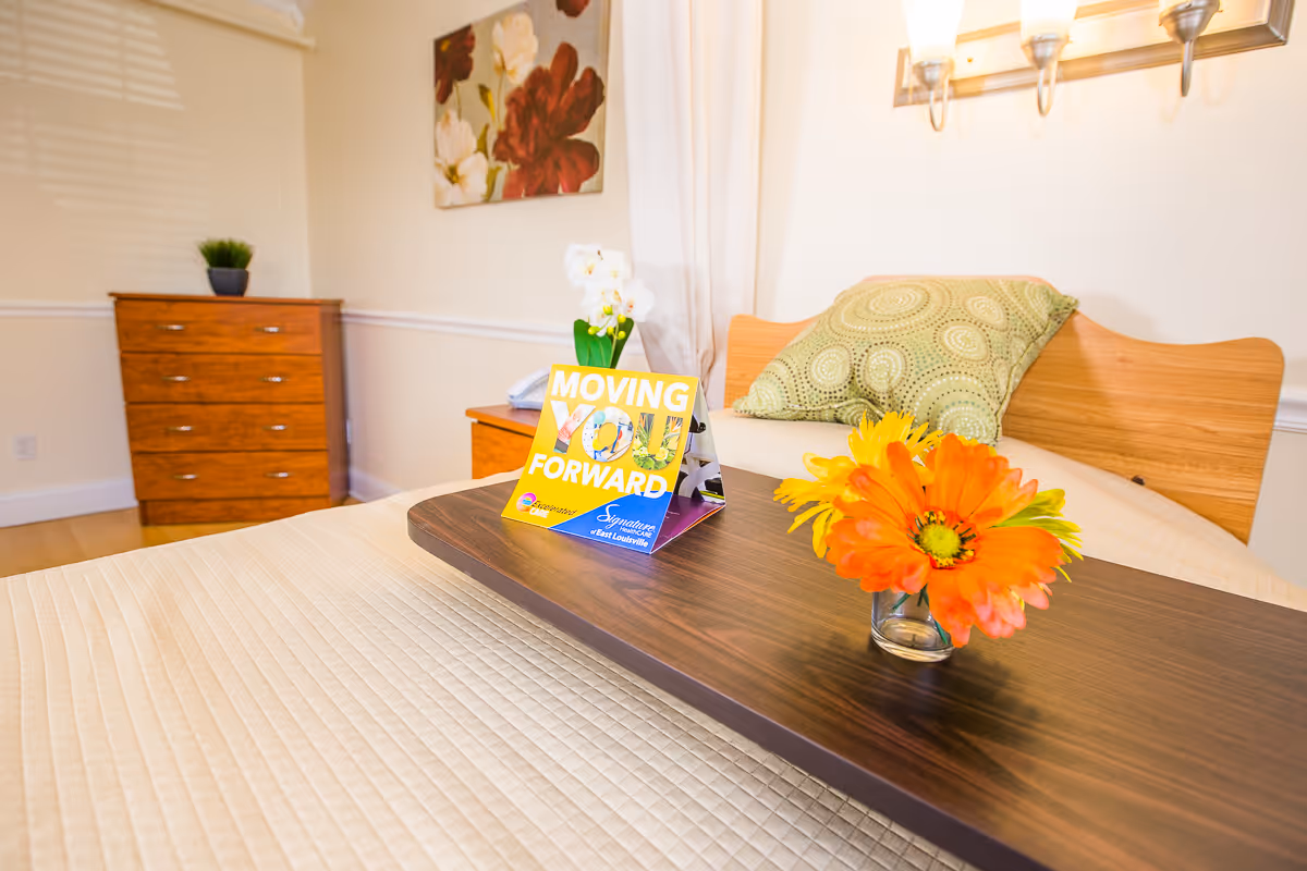 A neatly made bed with a wooden headboard and a green patterned pillow. On the bed is a dark wooden tray holding a small glass vase with bright orange and yellow flowers and a colorful sign that reads 'MOVING YOU FORWARD' with the Signature HealthCARE of East Louisville logo. In the background, there is a wooden dresser with a small potted plant on top and a floral painting on the wall.