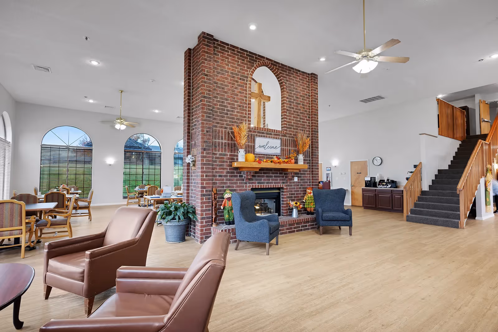 Spacious senior living common room with a central brick fireplace, multiple seating areas and dining tables, large arched windows, and a staircase.