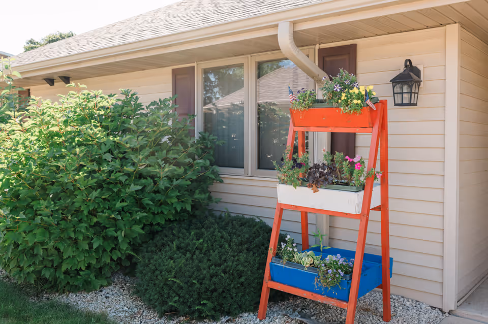 Exterior view of a beige-sided building with a window, shrubs, and a red ladder-style planter holding colorful flowers.