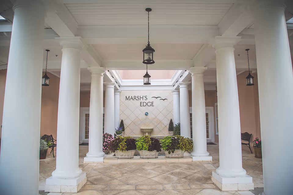 Entrance area of Marsh's Edge featuring large white columns, hanging lantern-style lights, potted plants, benches, and a wall with the facility name and decorative birds above a small water fountain.
