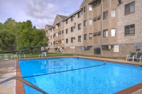 Outdoor swimming pool with clear blue water surrounded by a concrete deck and metal handrails. Several white lounge chairs and tables are placed around the pool area. In the background, there is a multi-story residential building with beige and brown exterior walls and multiple windows. Trees and greenery are visible on the left side under a partly cloudy sky.